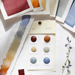 May include: A collection of watercolour supplies, including colour swatches, dot cards, and paint pans. The colour palette features shades of red, orange, yellow, grey, and blue. A small sprig of blue flowers and a paintbrush are also present.