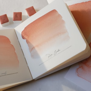 Può includere: Un set di acquerelli con un taccuino con campioni di colore rosa e pesca. Le pagine del taccuino mostrano sfumature di colore e i nomi "Cotton candy" e "Rose Ember". Sono visibili anche tre piccole pastiglie di vernice quadrate.