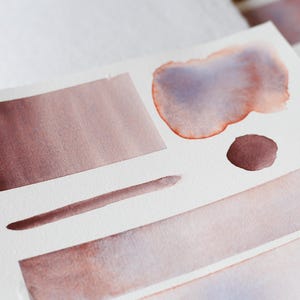 Könnte beinhalten: Weißes Aquarellpapier mit verschiedenen Farbfeldern in Rotbraun und Lila. Die Muster umfassen ein Rechteck, einen Kreis, eine lange Linie und eine große unregelmäßige Form, die Farbverläufe und Mischtechniken zeigen.