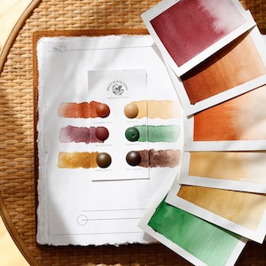 Könnte beinhalten: Aquarellfarbmuster in verschiedenen Rot-, Orange-, Grün- und Brauntönen auf weißem Papier. Das Papier trägt den Text "Hello Black Olive" und Farbbezeichnungen. Das Bild befindet sich auf einer gewebten Oberfläche.