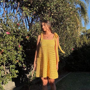 Peut inclure: Robe jaune au crochet avec encolure carr&eacute;e et bretelles nou&eacute;es. La robe arrive au genou et pr&eacute;sente un motif ajour&eacute; ample. La robe est faite d'un fil jaune vif et convient aux temp&eacute;ratures chaudes.