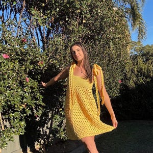 Peut inclure: Une robe jaune vif en crochet avec un d&eacute;collet&eacute; carr&eacute; et des bretelles nou&eacute;es aux &eacute;paules. La robe pr&eacute;sente un motif ajour&eacute;. La personne est debout &agrave; l'ext&eacute;rieur devant un buisson vert.