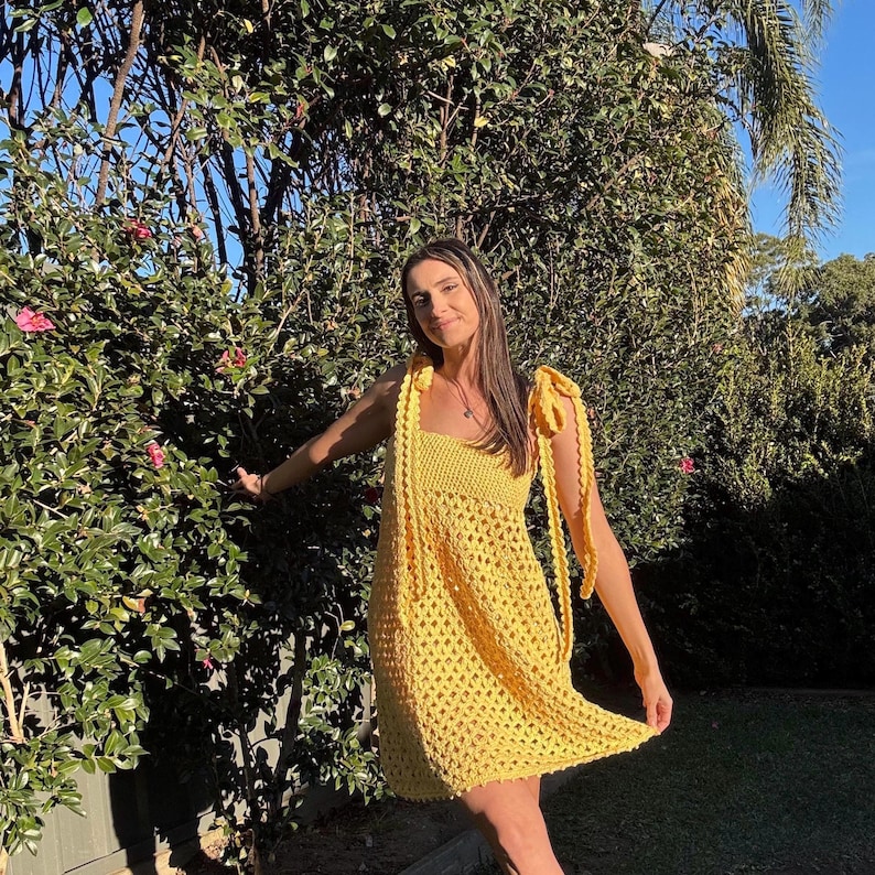 Peut inclure: Une robe jaune vif en crochet avec un d&eacute;collet&eacute; carr&eacute; et des bretelles nou&eacute;es aux &eacute;paules. La robe pr&eacute;sente un motif ajour&eacute;. La personne est debout &agrave; l'ext&eacute;rieur devant un buisson vert.