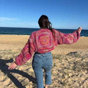 May include: A pink and red crochet bolero with long sleeves. The bolero has a square design on the back and sleeves, with a circular pattern in the centre. The garment is worn with blue jeans on a sandy beach.