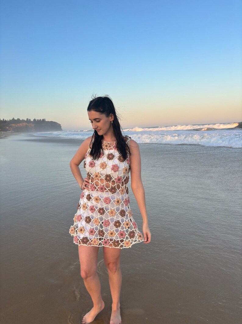 Girl in crochet flower dress standing at beach looking to left.