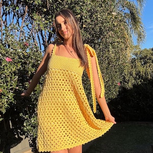 Peut inclure: Une robe jaune vif en crochet avec un col carr&eacute; et des bretelles nou&eacute;es. La robe pr&eacute;sente un motif ajour&eacute; textur&eacute; et une jupe l&eacute;g&egrave;rement &eacute;vas&eacute;e. Le mod&egrave;le se tient &agrave; l'ext&eacute;rieur devant un feuillage vert.