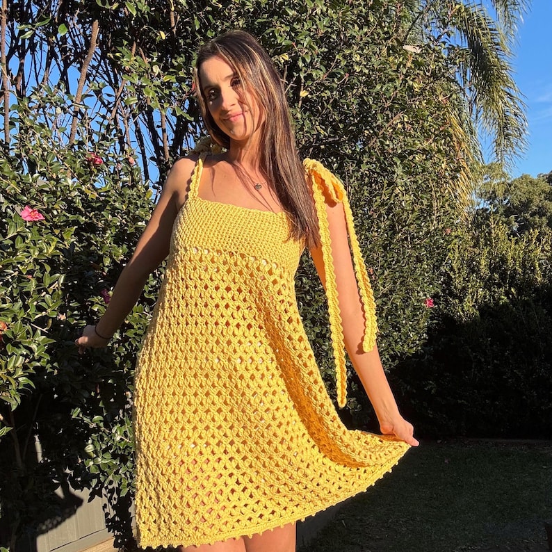 Peut inclure: Une robe jaune vif en crochet avec un col carr&eacute; et des bretelles nou&eacute;es. La robe pr&eacute;sente un motif ajour&eacute; textur&eacute; et une jupe l&eacute;g&egrave;rement &eacute;vas&eacute;e. Le mod&egrave;le se tient &agrave; l'ext&eacute;rieur devant un feuillage vert.