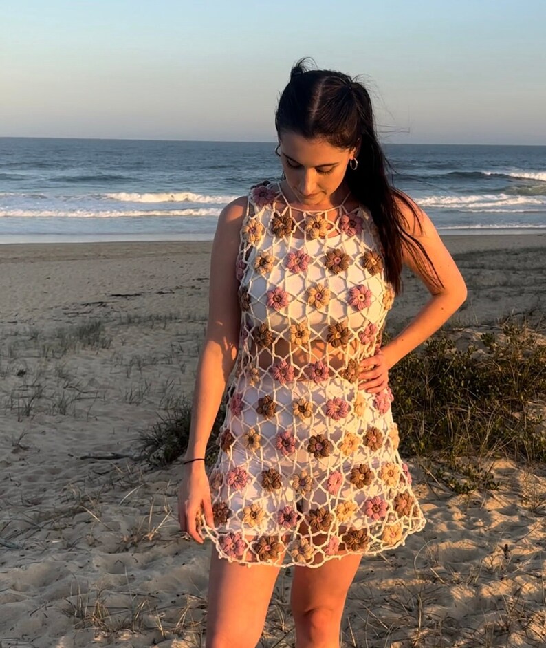 Girl in crochet flower dress standing at beach looking down.