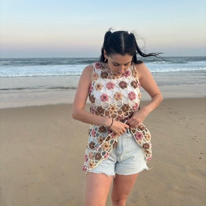 Girl in crochet flower dress standing at beach tucking dress into denim shorts and looking down.