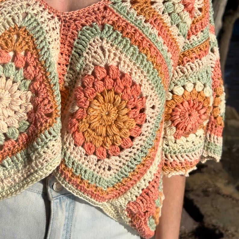 Pode incluir: Um top de croch&ecirc; com um padr&atilde;o geom&eacute;trico multicolorido em tons de p&ecirc;ssego, verde e creme. O top tem uma forma quadrada com bordas arredondadas e uma borda festonada.
