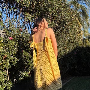 Peut inclure: Une robe en crochet jaune vif, &agrave; tissage ouvert, avec des bretelles spaghetti et des d&eacute;tails de n&oelig;uds. La robe arrive au genou et semble faite &agrave; la main. Le fond pr&eacute;sente un feuillage vert luxuriant et la lumi&egrave;re du soleil.