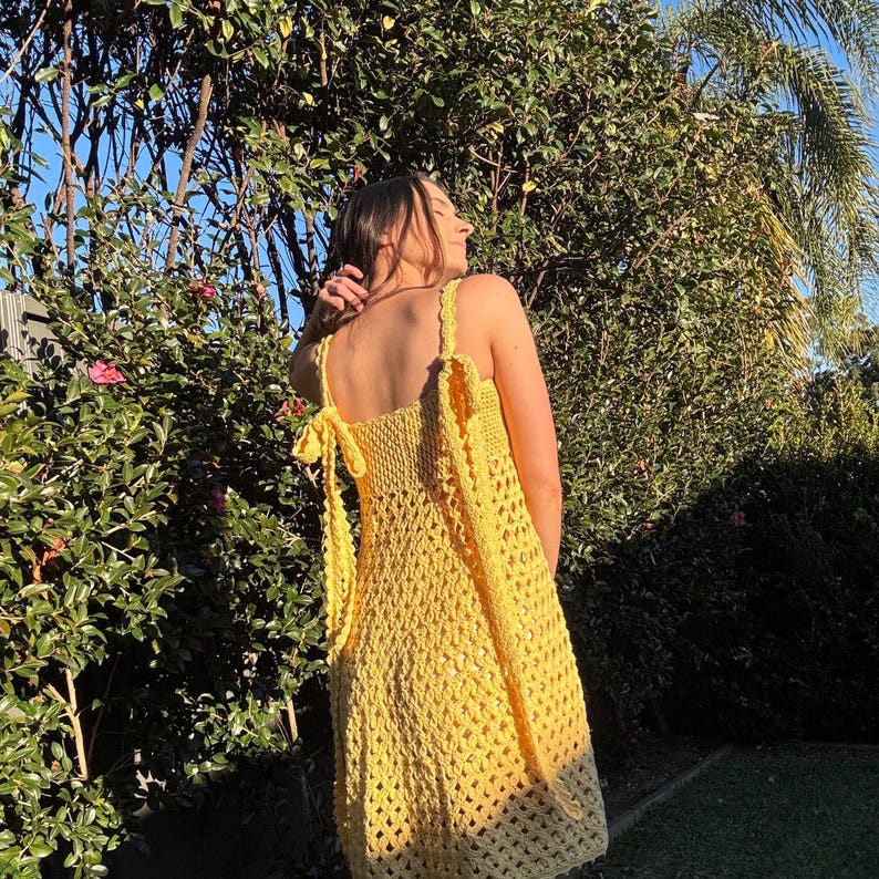 Peut inclure: Une robe en crochet jaune vif, &agrave; tissage ouvert, avec des bretelles spaghetti et des d&eacute;tails de n&oelig;uds. La robe arrive au genou et semble faite &agrave; la main. Le fond pr&eacute;sente un feuillage vert luxuriant et la lumi&egrave;re du soleil.