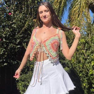 Può includere: Top all'uncinetto fatto a mano con un design con fiocco frontale. Il top presenta un motivo circolare con sfumature di arancione, verde e rosa. Le cinghie sono legate alle spalle. Il top è abbinato a una gonna bianca.