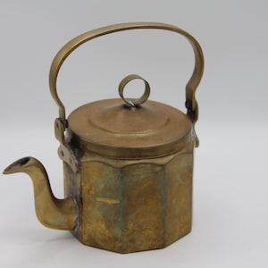 May include: A hexagonal brass teapot featuring a weathered, golden-brown patina. The vessel includes a curved spout, a flat lid with a small circular handle, and a tall, arched swing handle attached to the sides with metal rivets.