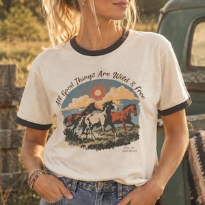 May include: A cream-colored ringer t-shirt with dark trim on the collar and sleeves. The graphic features three horses running across a landscape under a sun and clouds, with the text "All Good Things Are Wild & Free" and "Save our wild horses."