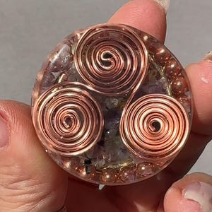 Op de afbeelding: Een ronde, heldere harsbol met drie ingebedde koperen draadspiralen. De bol is gevuld met kleine, roze en witte kristallen.