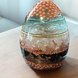 Puede incluir: Una esfera de vidrio transparente con capas de cuentas de cobre, líquido transparente, cristales blancos y piedras negras. La esfera tiene forma de huevo y está sobre una superficie de madera.