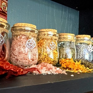Może przedstawiać: Rząd szklanych słoików z korkowymi pokrywkami i sznurkowymi akcentami, każdy zawierający różne suszone zioła i przyprawy. Każdy słoik ma srebrny wisiorek pentagramu wiszący z pokrywki.