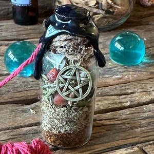 May include: A glass bottle with a black wax seal and a pentagram charm inside. The bottle is filled with dried herbs and crystals. The bottle is sitting on a wooden surface with a red string and a blue glass orb.