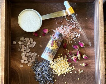 Op de afbeelding: Een houten dienblad gevuld met ingrediënten voor een spa-behandeling. Inclusief een maatlepel met witte korrels, een kleine glazen fles met gedroogde bloemen, rozenkwarts stenen, lavendel en andere natuurlijke elementen.