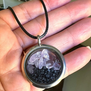 May include: A silver-colored pendant necklace with a round glass locket. The locket contains light purple stones and black gravel. The necklace has a black cord.