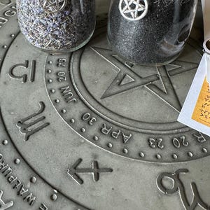 Può includere: Due barattoli di vetro con ciondoli a pentagramma in cima, riempiti di erbe nere e viola, appoggiati su una superficie circolare grigia con un design a ruota zodiacale e il testo "MAY EVE WALPURG" e "APR 30 JUN 20 23".