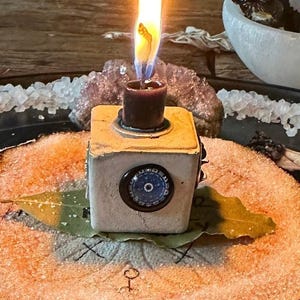 May include: A small white ceramic candle holder with a blue and white eye design on the front. The candle holder is lit and the flame is casting a warm glow on the surrounding area. The candle holder is sitting on a wooden surface with a bay leaf and white crystals.