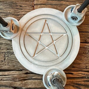 Può includere: Un altare circolare in cemento grigio chiaro con un disegno a pentagramma. L'altare ha tre supporti attaccati con aste di metallo nero. L'altare è posto su una superficie di legno stagionato.