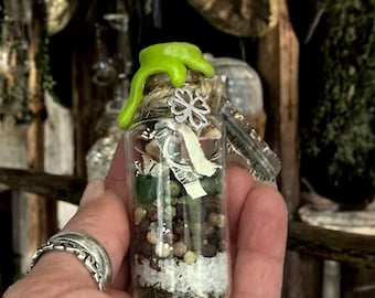 May include: A small glass jar filled with various herbs, spices, and crystals. The jar has a green, dripping wax seal and a silver charm with a four-leaf clover design.