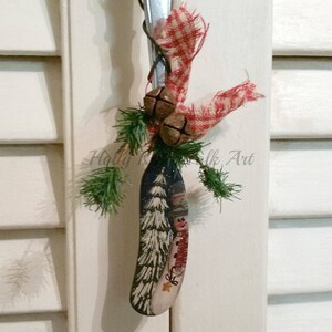 May include: A painted wooden spoon ornament with a snowman design and a green pine sprig. The spoon is decorated with a red and white gingham ribbon and jingle bells.