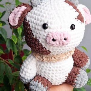 May include: A crocheted cow plush toy, predominantly white with brown patches and grey hooves. It features pink inner ears, a pink nose, and black safety eyes. A tan scarf encircles its neck. The soft toy is designed to be cuddled.