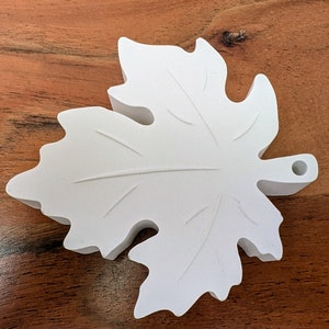 Könnte beinhalten: Ein weißer Keramik-Blatt-Anhänger mit einem Loch oben zum Aufhängen. Das Blatt hat ein detailliertes Adernmuster und eine leicht strukturierte Oberfläche.