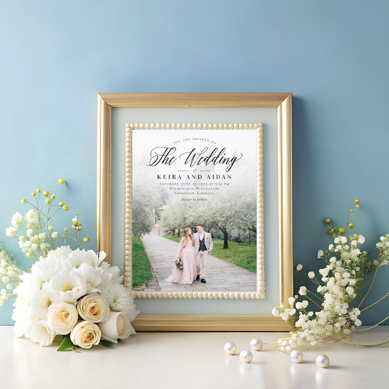 Puede incluir: Una invitaci&oacute;n de boda con un marco dorado y un fondo blanco. El texto dice "Usted est&aacute; invitado a La boda de Keira y Aidan S&aacute;bado, 22 de junio de 2025 a las 5:30 PM Wilmington Plantation Savannah, Georgia Cena a seguir". Una foto de la pareja caminando por un sendero est&aacute; en el centro de la invitaci&oacute;n.