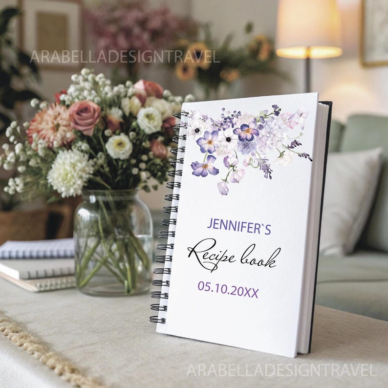 May include: A white spiral bound recipe book with a watercolor floral design in shades of purple and white. The text on the cover reads "Jennifer's Recipe Book 05.10.20XX".