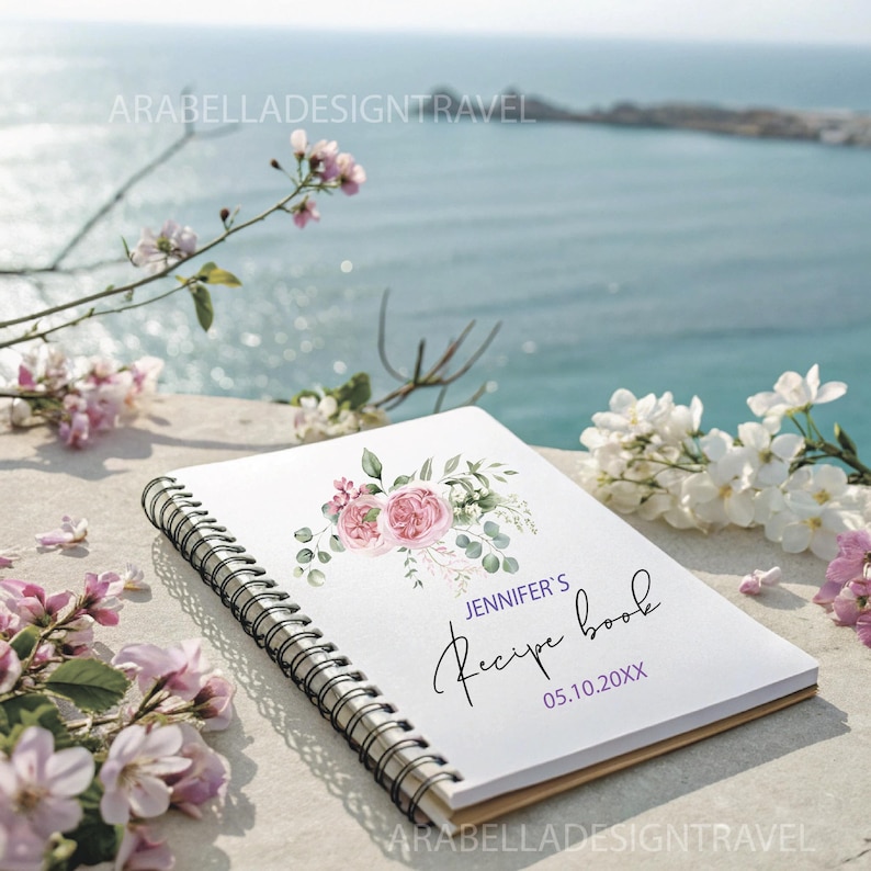May include: A white spiral bound recipe book with a watercolor floral design featuring pink roses and green leaves. The text "Jennifer's Recipe Book" is written in black cursive with the date "05.10.20XX" below it.