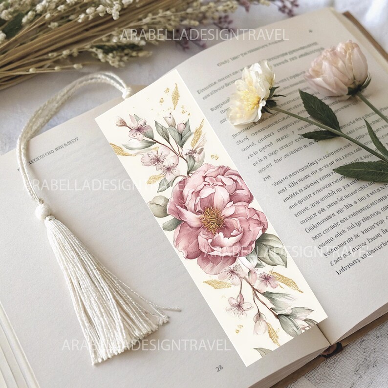 May include: A floral bookmark featuring a large pink peony and delicate white flowers, accented with green leaves and gold details. The bookmark has a white tassel and the text "ARABELLADESIGNTRAVEL". It rests on the open pages of a book.