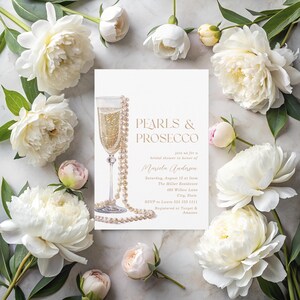 Puede incluir: Una tarjeta de invitaci&oacute;n blanca con letras doradas para una despedida de soltera. La invitaci&oacute;n dice "Pearls & Prosecco" e incluye el nombre de la novia, la fecha, la hora y el lugar. La tarjeta presenta una ilustraci&oacute;n de acuarela de una copa de champ&aacute;n con un collar de perlas.