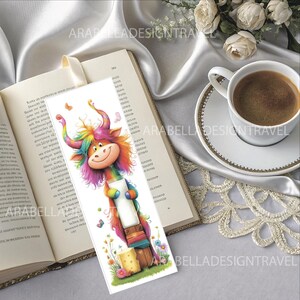 May include: A colorful bookmark featuring a cartoon character with rainbow-colored hair and horns, holding a paint roller. The bookmark is placed in an open book with a cream-colored ribbon. A cup of coffee sits on a saucer nearby.