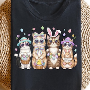 May include: Black t-shirt with a colourful graphic of four cats celebrating Easter. The cats wear sunglasses, hats, and bows. They hold Easter baskets and eggs, with the text "HAPPY EASTER" on a sign held by one cat. The design is set against a black background.