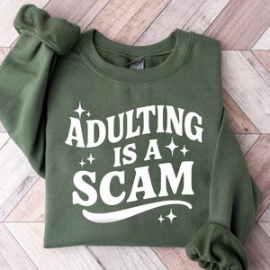 May include: An olive green sweatshirt with the phrase "Adulting is a Scam" in white, stylized text. The sweatshirt is folded, with the sleeves cuffed. The background is a light-colored wooden surface.