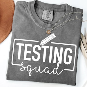 Può includere: Una maglietta grigia con le parole "TESTING squad" in bianco e un disegno di una matita. Una collana d'oro con un ciondolo è drappeggiata sulla maglietta. Un cappello marrone e una pianta secca sono sullo sfondo.