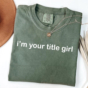 May include: An olive green t-shirt with the phrase "i'm your title girl" printed in white. A gold necklace with a circular pendant rests on the shirt. The shirt is folded and displayed with a brown hat and dried plant.