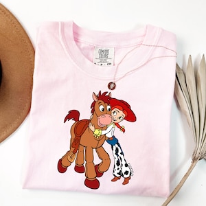 May include: A light pink Comfort Colors t-shirt with a cartoon graphic of a cowgirl hugging a horse. The cowgirl wears a red hat, white shirt, blue jeans, and black and white chaps. The horse is brown with a saddle. A gold necklace is on the shirt.