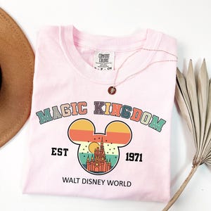 May include: A light pink t-shirt with the words "MAGIC KINGDOM" in colorful letters above a Mickey Mouse head design. The design includes a castle and the text "EST 1971 WALT DISNEY WORLD". A gold necklace is on the shirt.
