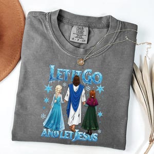 May include: Gray Comfort Colors t-shirt featuring the phrase "Let It Go and Let Jesus" with an illustration of Jesus and two characters. The shirt has a crew neck and short sleeves. A silver necklace with a pendant is on the shirt.