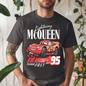 May include: Dark gray t-shirt featuring Lightning McQueen from the Cars movie. The design includes the text "Lightning McQueen", a red race car, the number "95", and the words "Think Fast".