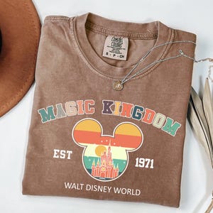 May include: A brown t-shirt with the words "MAGIC KINGDOM" in colorful letters and a Mickey Mouse head design with a castle and sunset graphic. The shirt also has "EST 1971" and "WALT DISNEY WORLD" printed on it.