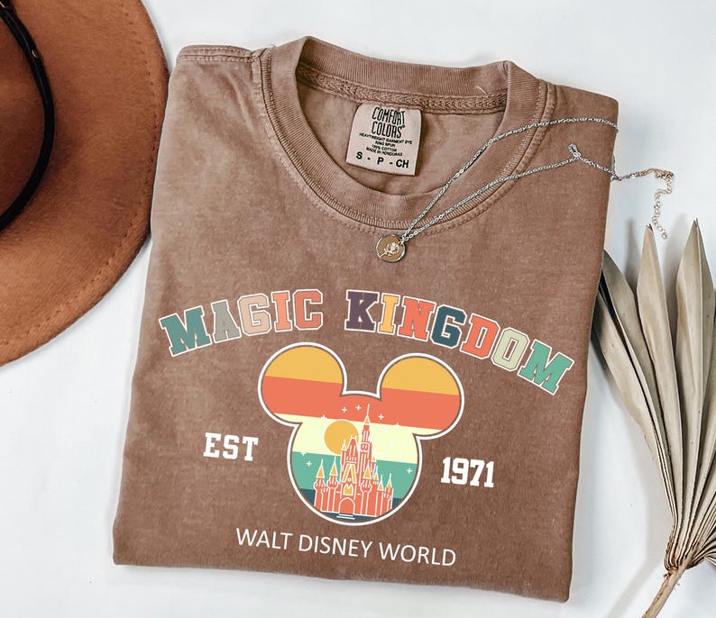 May include: A brown t-shirt with the words "MAGIC KINGDOM" in colorful letters and a Mickey Mouse head design with a castle and sunset graphic. The shirt also has "EST 1971" and "WALT DISNEY WORLD" printed on it.