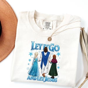 May include: Cream-colored t-shirt with the text "Let It Go and Let Jesus" in blue. The graphic features Jesus holding hands with two cartoon characters. A gold necklace and a brown hat are also visible.