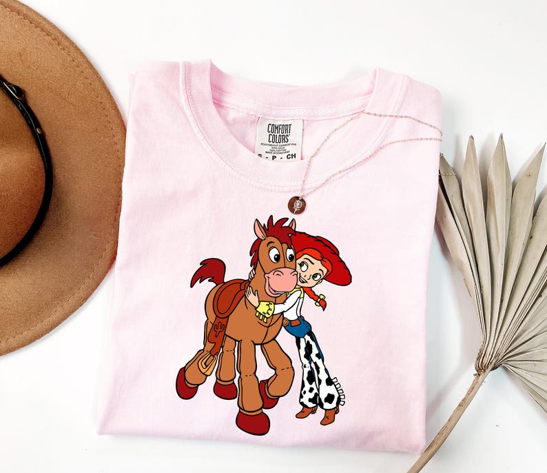 May include: A light pink Comfort Colors t-shirt with a cartoon graphic of a cowgirl hugging a horse. The cowgirl wears a red hat, white shirt, blue jeans, and black and white chaps. The horse is brown with a saddle. A gold necklace is on the shirt.